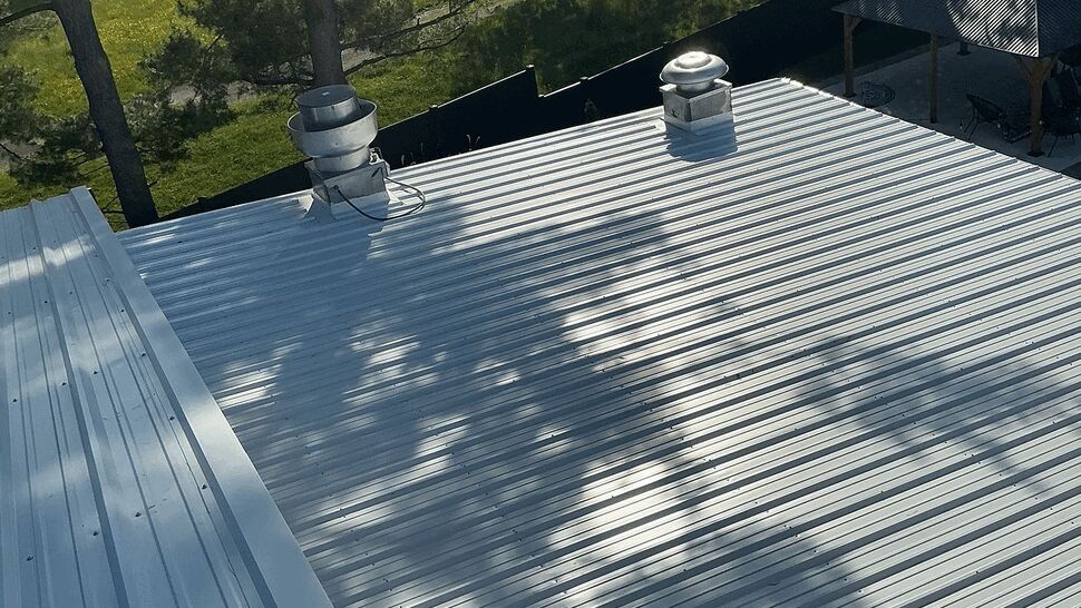 Close view of corrugated metal roof panels with roof vents and tree shadows.