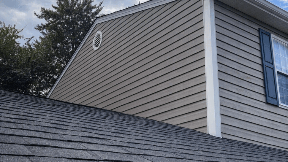 Siding Roof and Gutter Detail | Tycos Roofing And Siding Inc Gray house siding above dark shingles with white gutter and trim under a blue sky