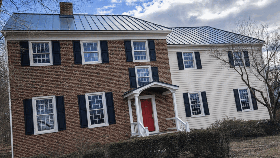Standing-seam metal roof on colonial | Tycos Roofing And Siding Inc Front view of brick and siding colonial with standing-seam metal roof and a red front door