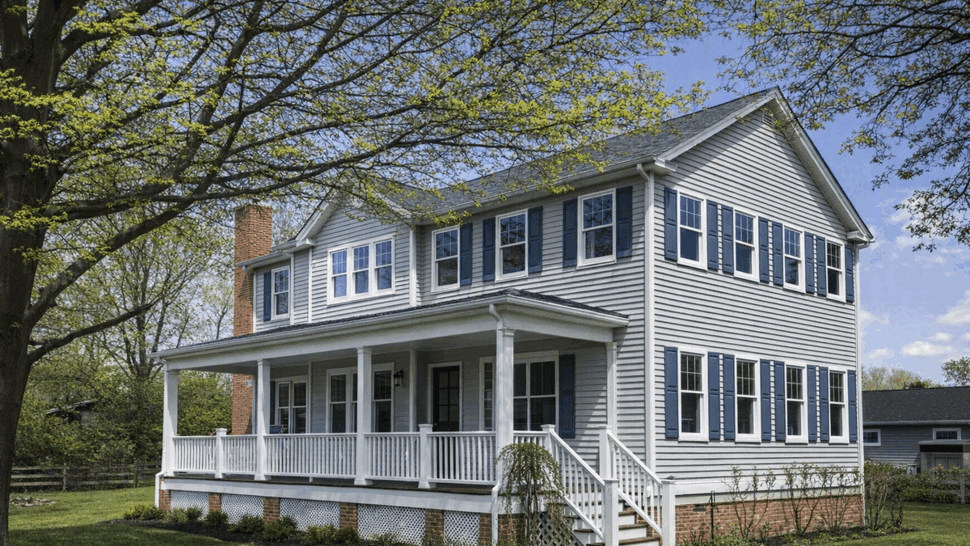 Two-story house with gray siding, wraparound porch and white trim under leafy trees.