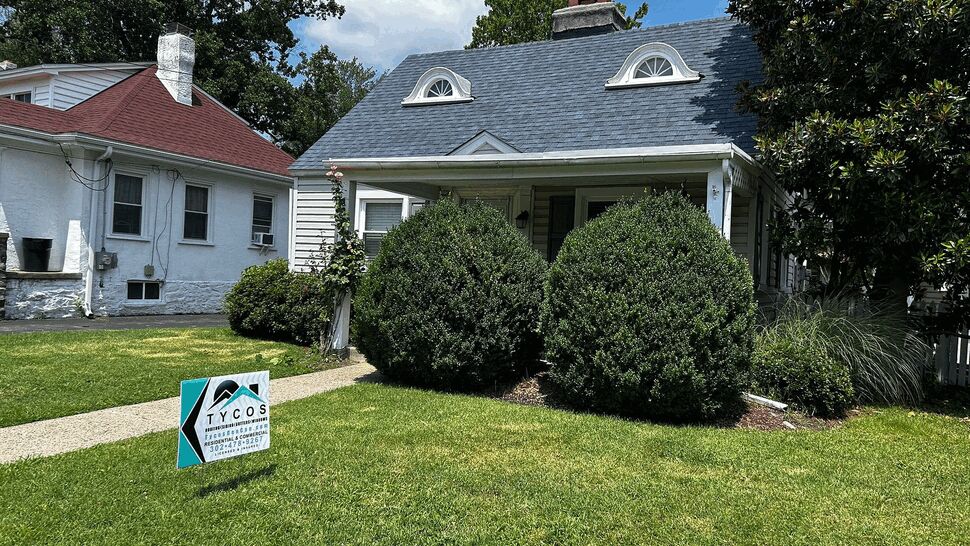 Front yard of a cape cod house with a Tycos sign on the lawn and a gray shingle roof.