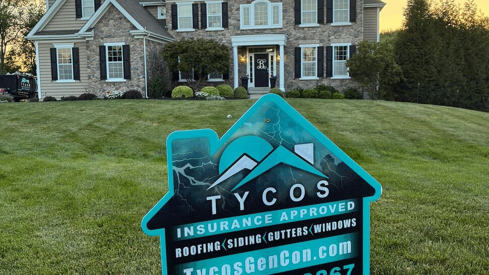 Yard sign advertising Tycos Roofing And Siding in front of a large home at sunset