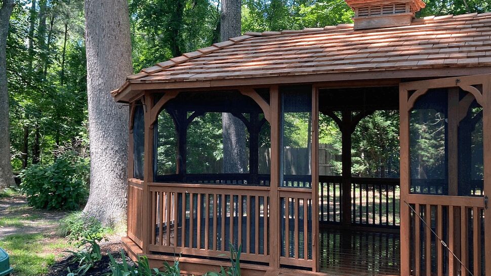 Gazebo side profile with landscaping | Tycos Roofing And Siding Inc Side view of a gazebo next to large trees and landscaped plantings.