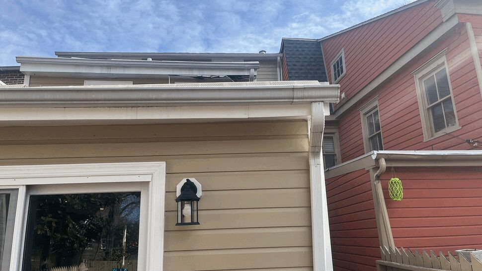 White gutter corner and downspout attached to beige siding between houses.