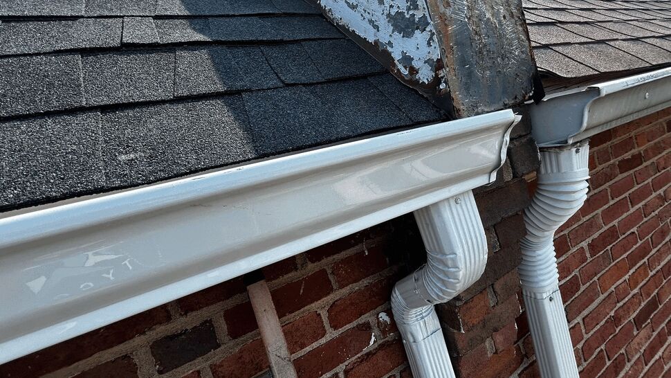 Close-up of white gutter, elbow and downspout against brick and shingles.