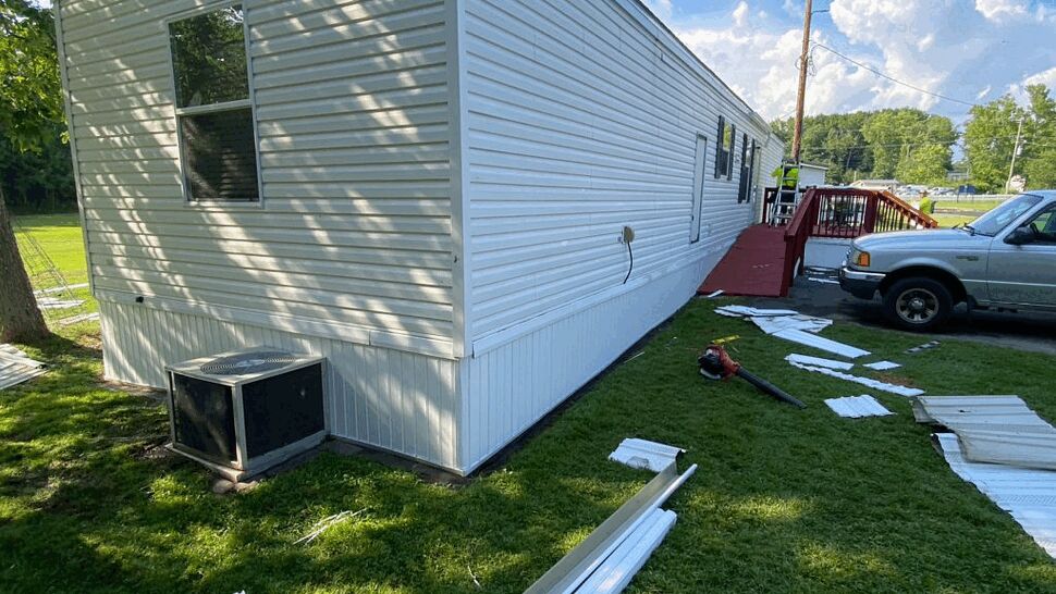 Side view of a white mobile home with vinyl siding, skirting, and replacement panels on the lawn.