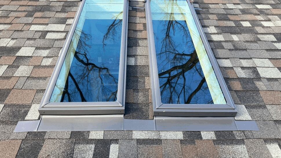 Paired skylights on shingled roof | Tycos Roofing And Siding Inc Two rectangular skylights installed side by side on a shingled roof reflecting tree branches.