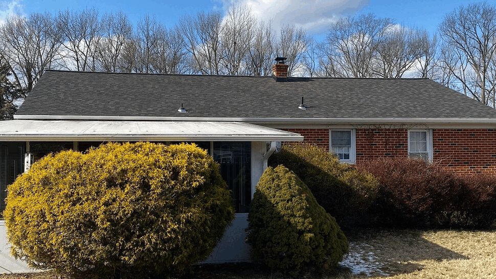 Brick ranch house with dark asphalt shingle roof under a blue sky and clouds.