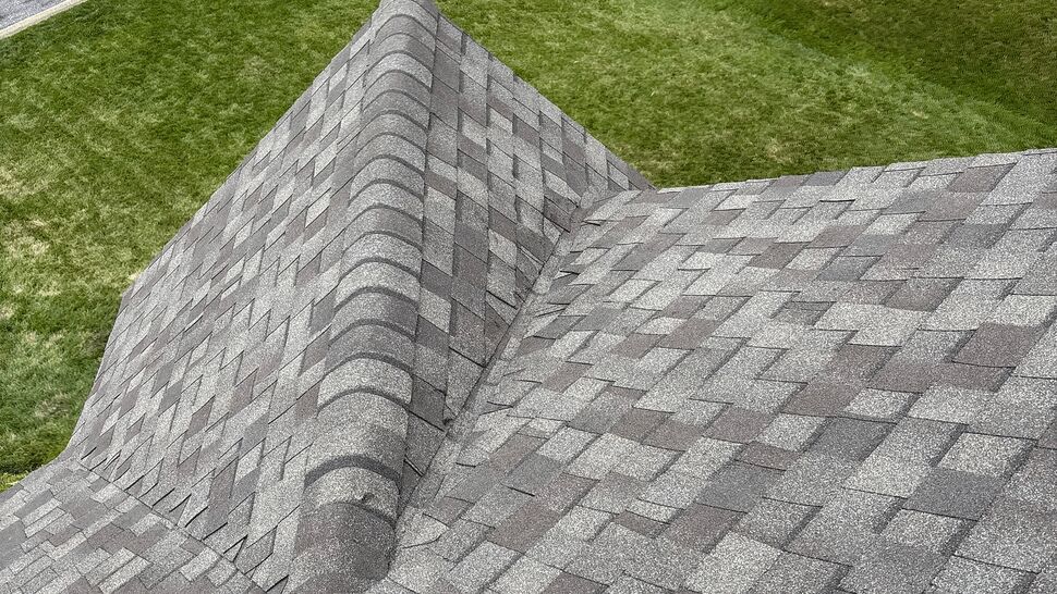 Close-up of asphalt shingle roof valley and ridge with company debris trailer visible below.