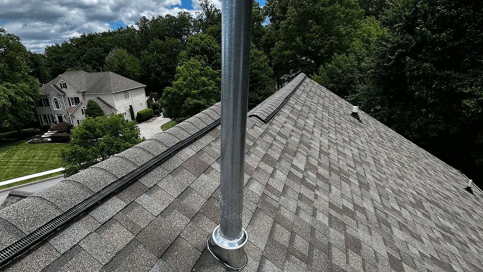 Metal vent pipe penetrating gray asphalt shingle roof under a partly cloudy sky