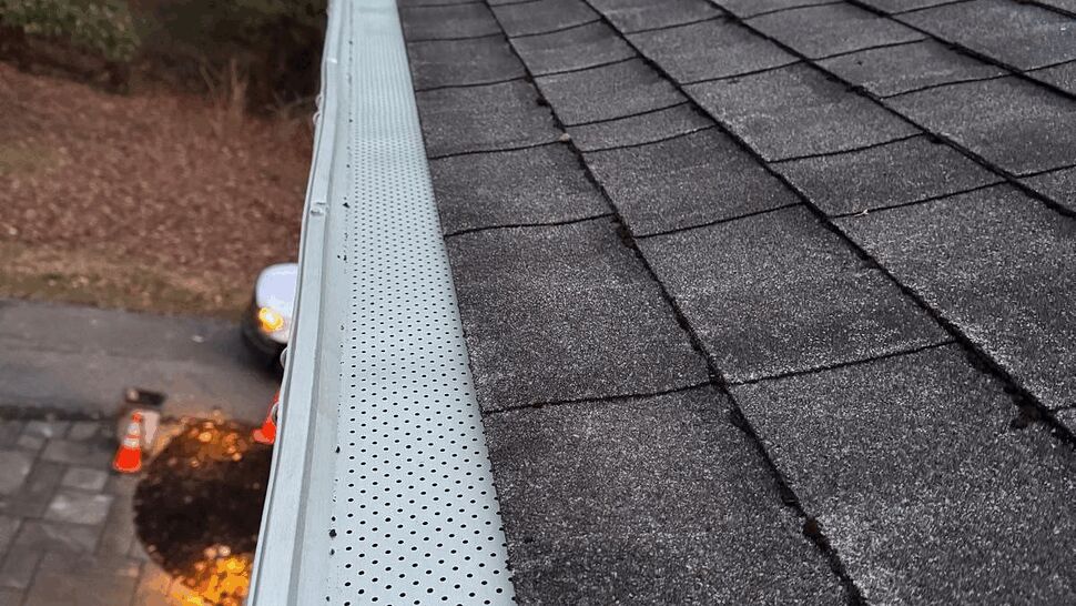 Close-up of asphalt shingle roof and eave from above looking toward treeline