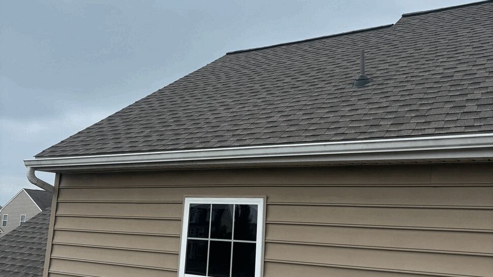 Roof slope with white gutter above a second-story window on beige siding.