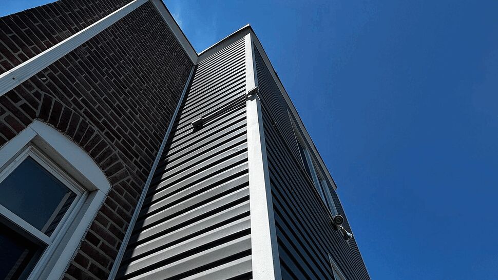 Siding and roofline detail | Tycos Roofing And Siding Inc Low-angle view of house corner showing gray siding and roofline under clear sky