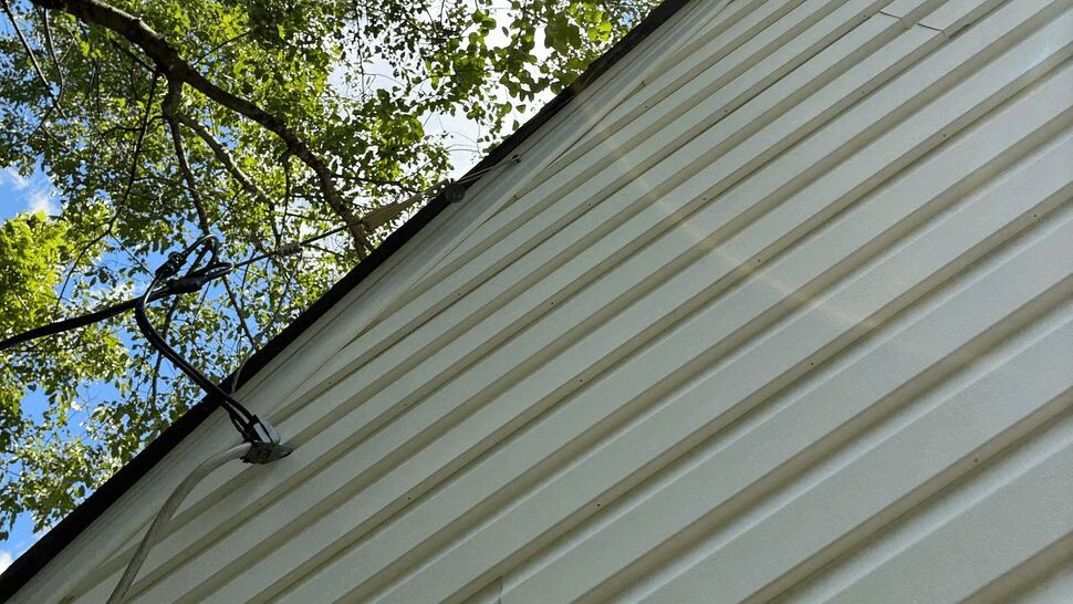 Siding and roofline beneath tree canopy | Tycos Roofing And Siding Inc Exterior siding and roofline beneath a tree canopy with utility cable visible