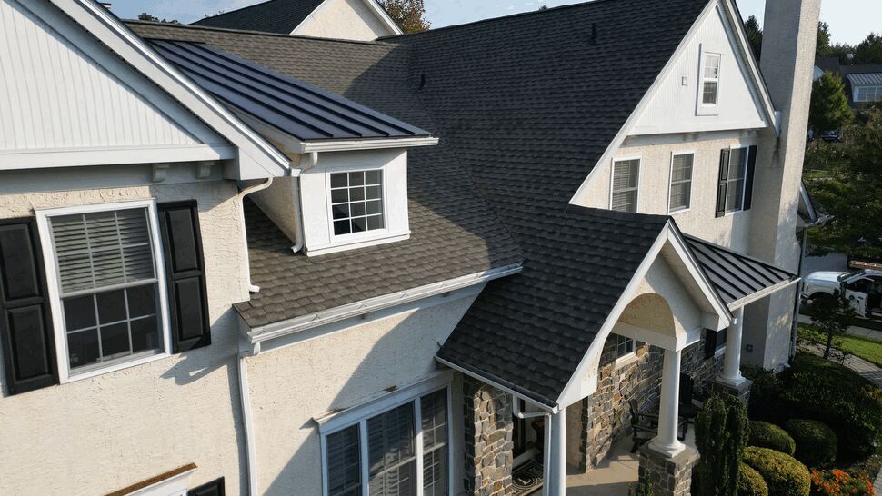 Aerial front view of a house with shingled roof, stone porch, columns and white siding.