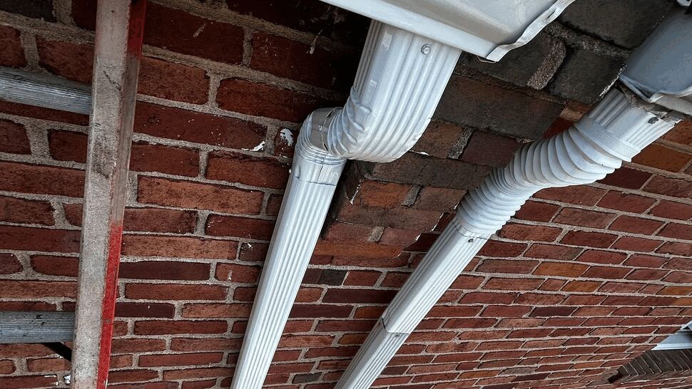 Two vertical downspouts fastened to a brick exterior beside a ladder.