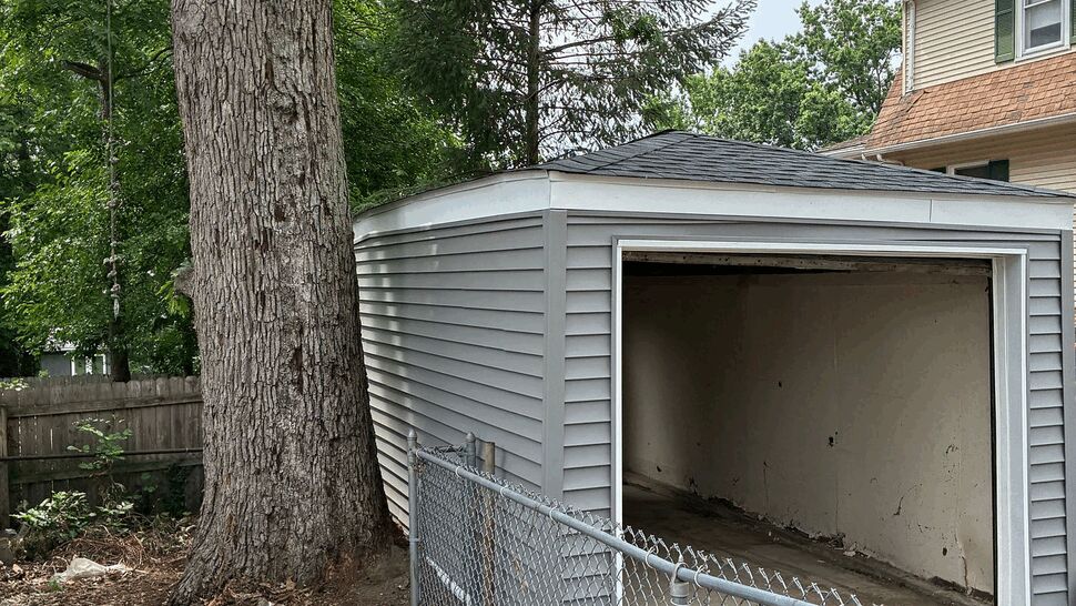 Small gray vinyl-sided garage with open bay door beside a large tree