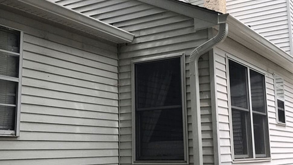 Corner of a white vinyl sided house with a downspout beneath tree branches.