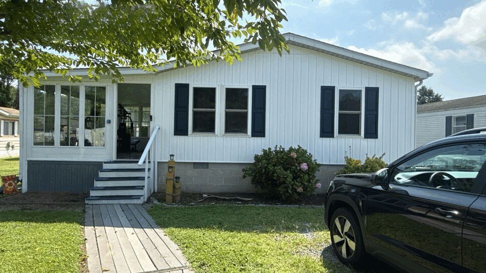 White-sided manufactured home with dark shutters, porch, and walkway