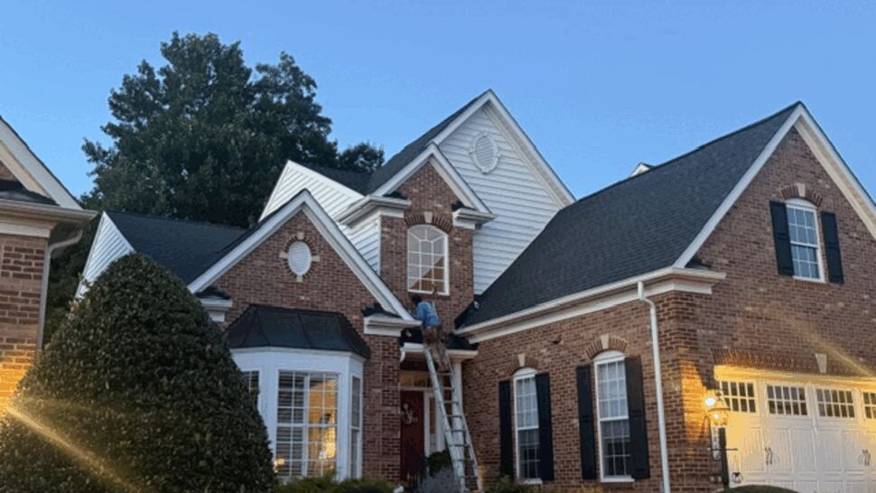 Brick-front house with ladder and crew replacing roof at dusk.