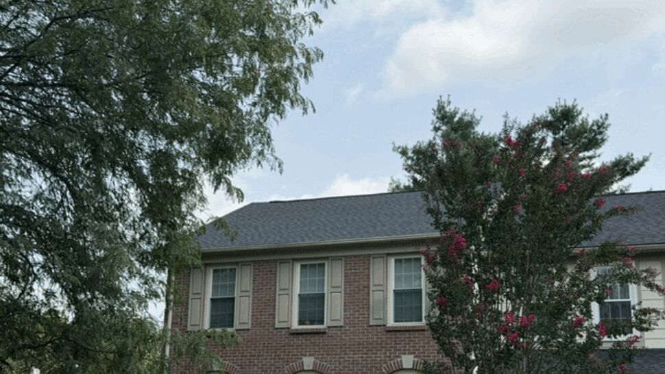 Two-story brick townhouse with dark shingle roof and trees in foreground
