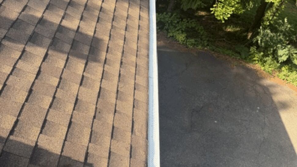 Close-up of brown asphalt shingles and white gutter along roof edge.