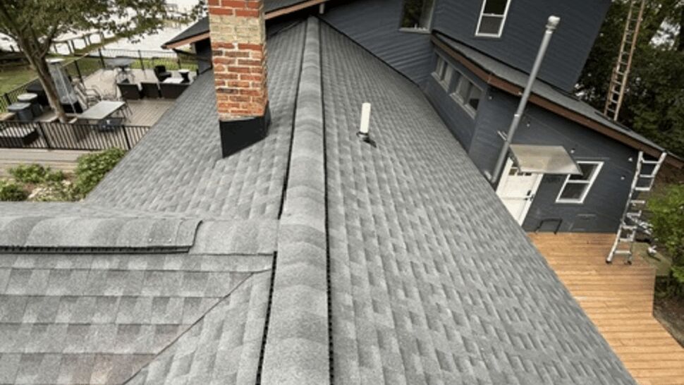 Aerial view of a gray shingled roof ridge with a brick chimney and surrounding trees.