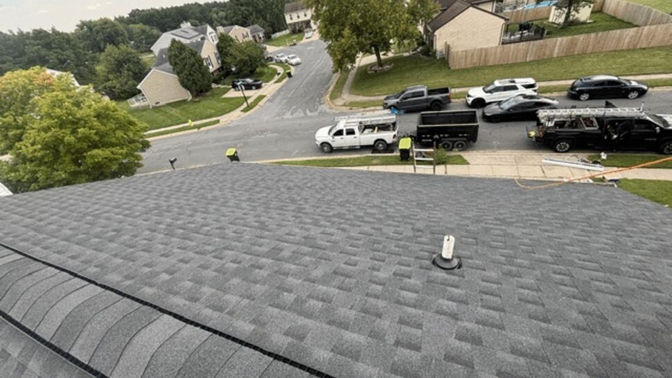 Close-up of a new gray asphalt shingle roof with a vent pipe and neighborhood view.