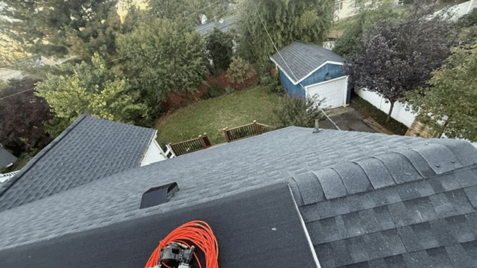 Gray shingle roof with ridge caps and roofing equipment overlooking a backyard.