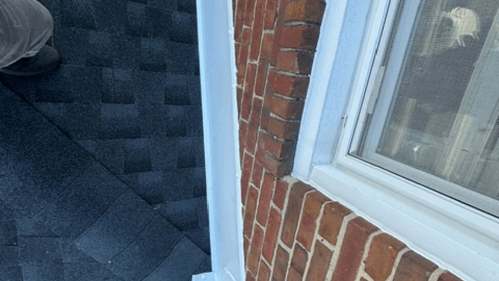 Roof Valley and Flashing Detail | Atlas Exteriors Close-up of gray shingles and white flashing beside a brick wall and window.