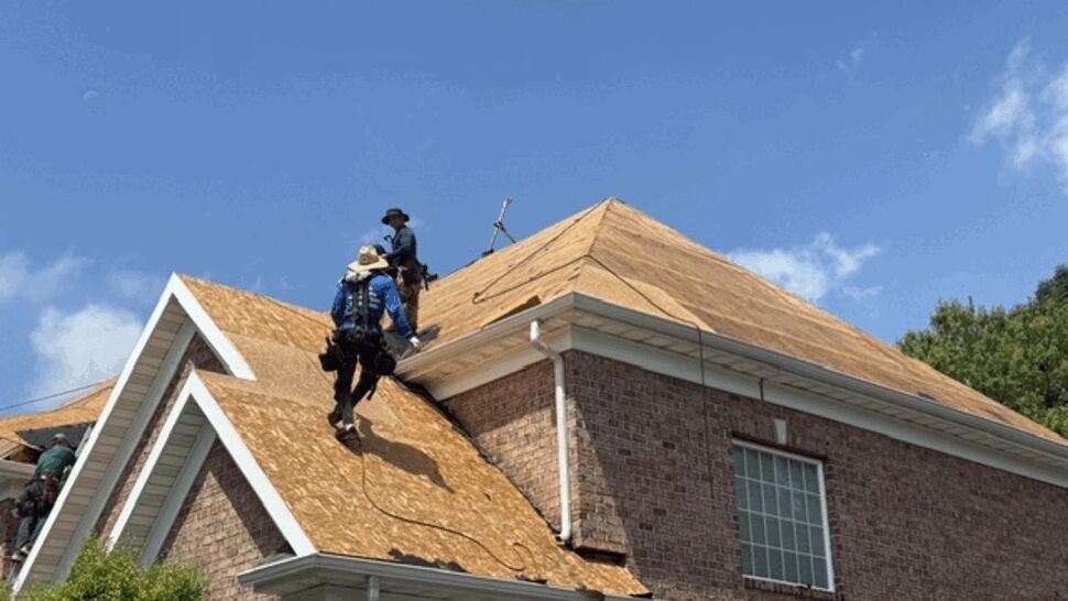 Workers installing plywood decking on a home roof under a clear sky.