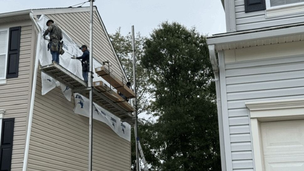 Two workers on scaffold fitting house wrap and panels near roofline.
