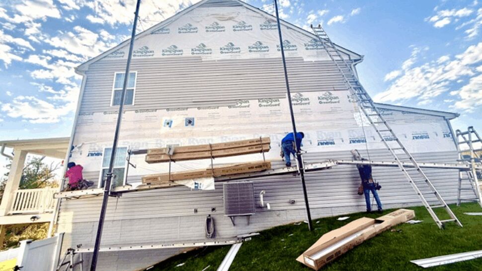 Workers installing light gray siding on a two-story home with scaffolding