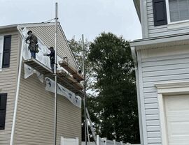 Two workers on scaffold fitting house wrap and panels near roofline.