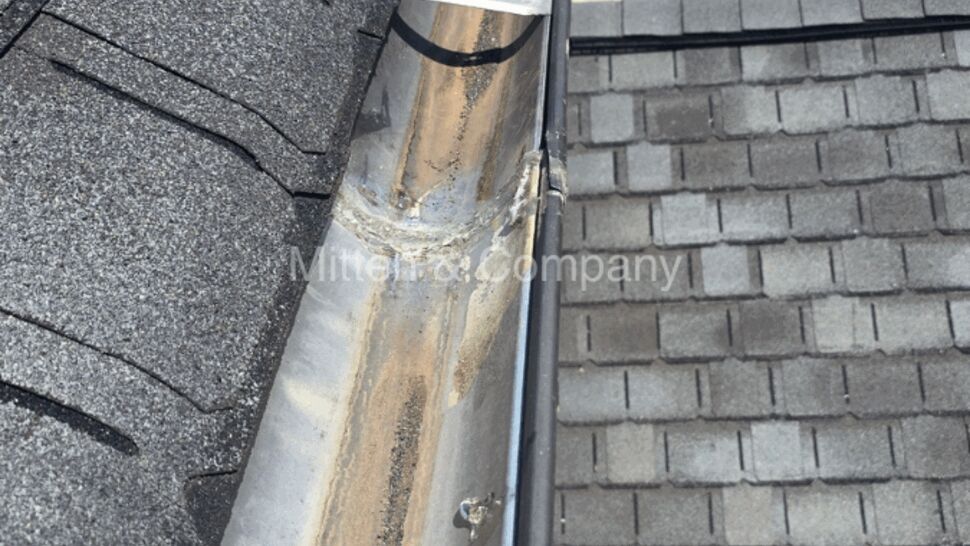 Top-down view of a metal gutter seam with dirt and adjacent asphalt roof shingles.