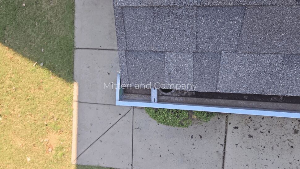 Roof Gutter Corner Inspection | Mitten & Company Close-up of a shingle roof edge and gutter beside a concrete walkway