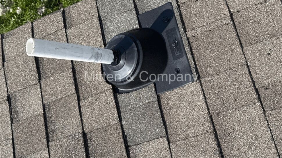 Black rubber vent boot with white pipe on shingled roof near greenery
