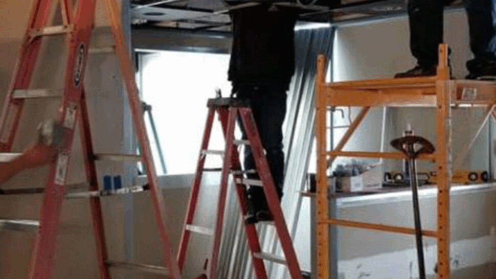 Workers on ladders and scaffolding installing a ceiling in an interior space.