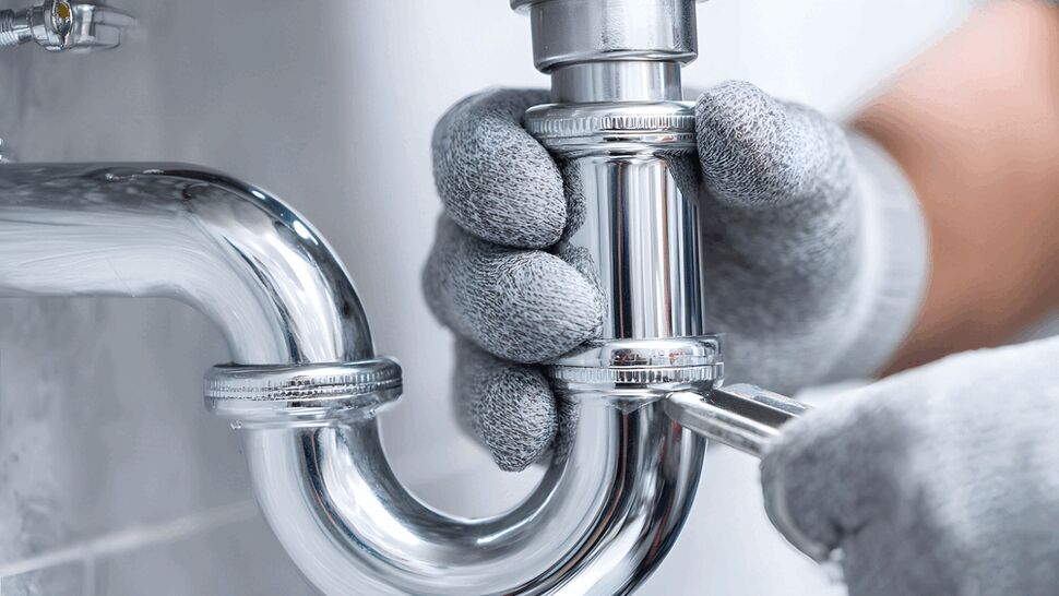 Gloved hands tightening a chrome P-trap under a sink with a screwdriver.
