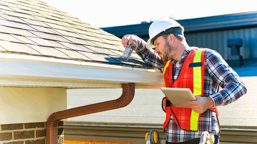 Worker on ladder inspecting gutter and roof while holding a tablet