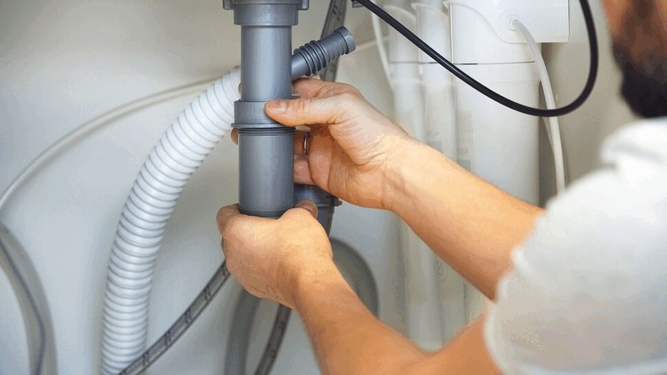 Hands fitting gray plastic drain pipe connections beneath a sink with hoses nearby.