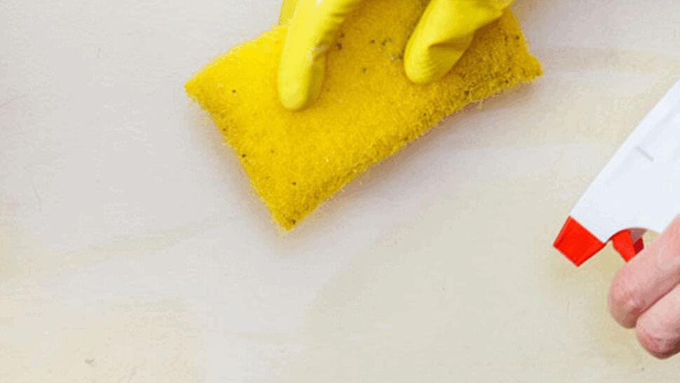 Gloved hand scrubbing a baseboard area with a sponge and spray cleaner to remove stains