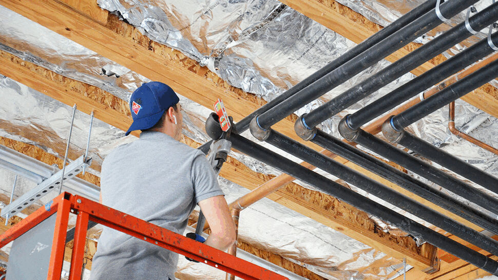 Worker on a lift installing black plumbing pipes against foil-insulated ceiling joists.