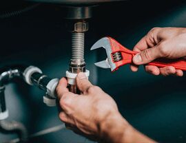 Hands using a red adjustable wrench to tighten piping beneath a sink.