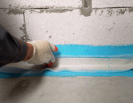 Worker applying blue waterproof membrane at concrete wall-floor joint