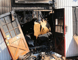 Open double doors revealing a charred interior and scattered debris at a damaged entrance.