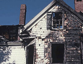 Two-story house exterior with extensive charring on siding and broken windows from a fire.