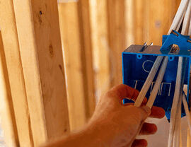 Hand guiding electrical cables into a blue outlet box in a wooden stud wall.