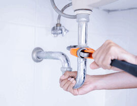 Technician using an orange pipe wrench on a metal P-trap under a sink.