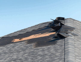 Ridge area with peeled back shingles and exposed roof decking under clear sky.